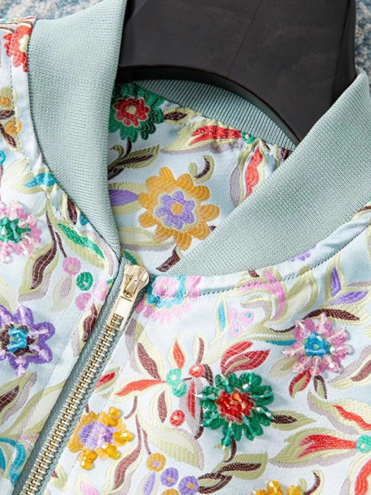 Close-up of floral embroidered bomber jacket with sequins, featuring a light blue fabric and ribbed collar. | SuzzaneMarket