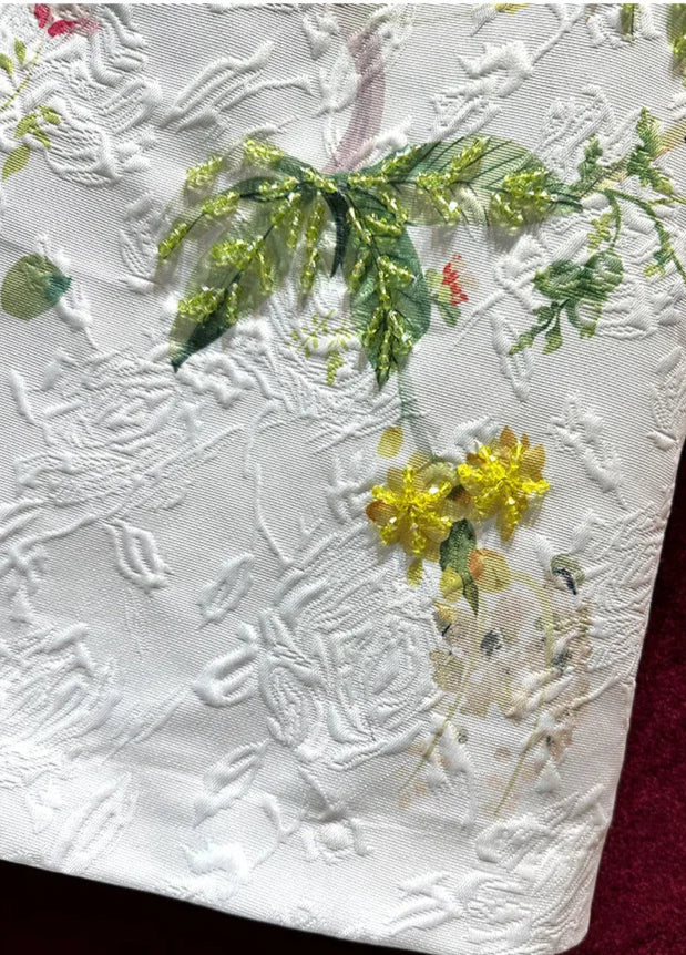 Close-up of embroidered floral design in white fabric, featuring yellow flowers and green leaves | SuzzaneMarket.