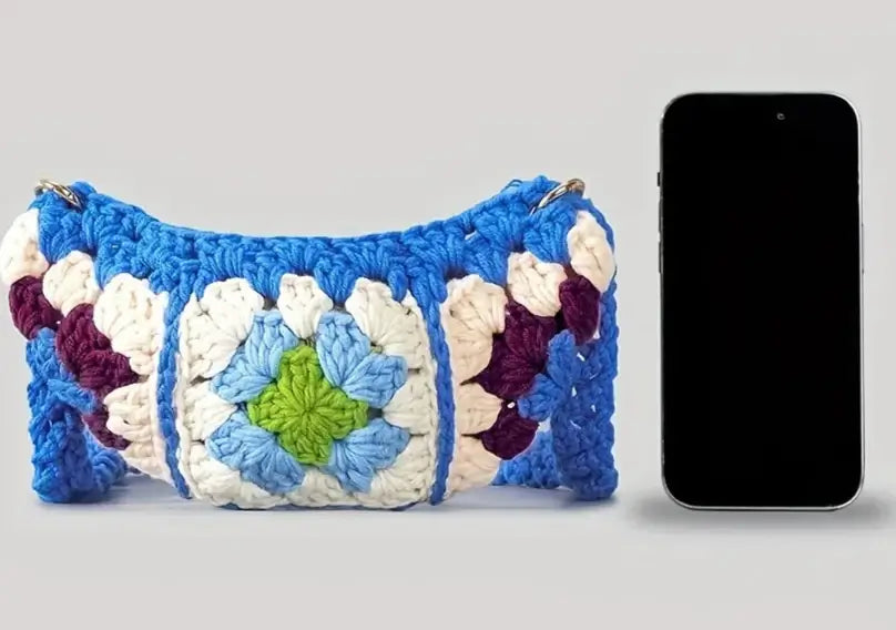 Handcrafted crochet banane bag in blue, featuring colorful patches, alongside a smartphone for size reference.